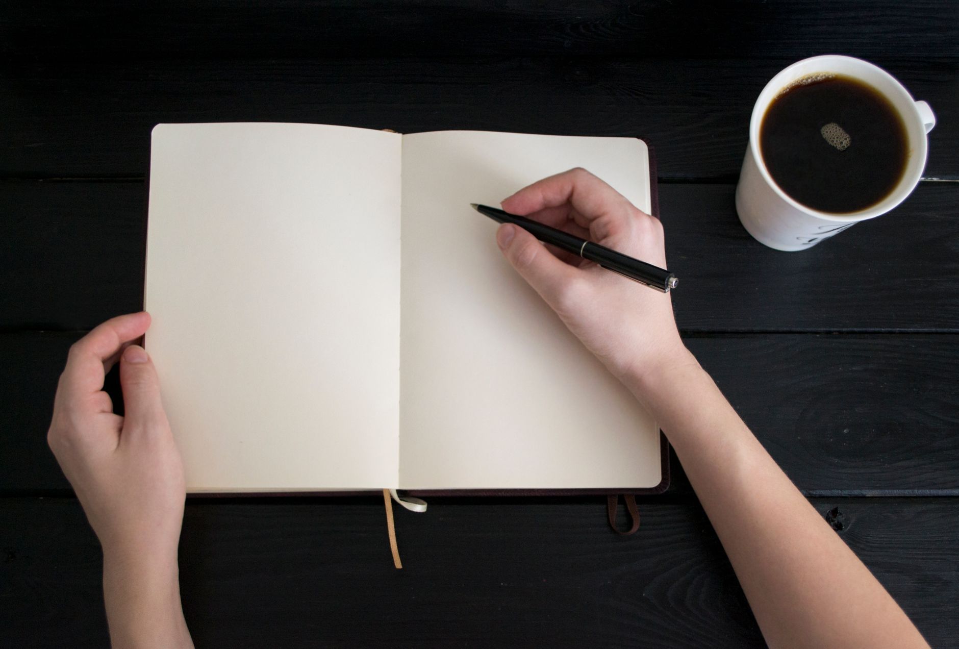 A hand writing in a notebook next to a cup of coffee on a wooden desk